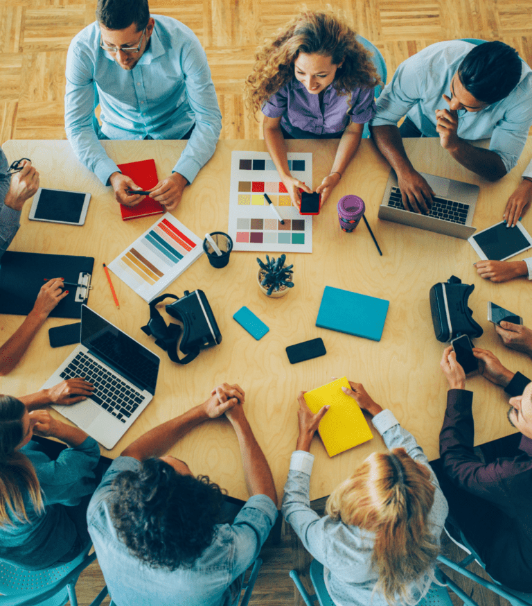 A group of people sitting around a boardroom table talking with laptops, notebooks and charts on the table in front of them