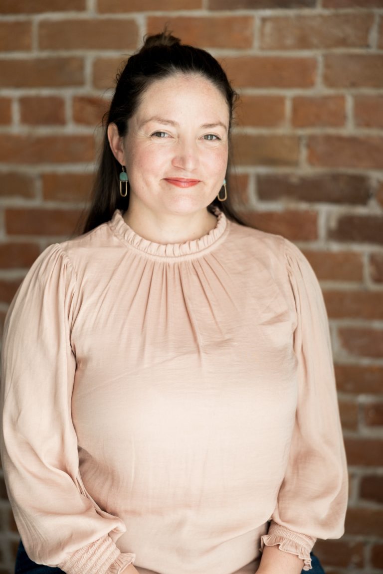 A smiling white woman with dark brown hair in a ponytail wearing a pink blouse.