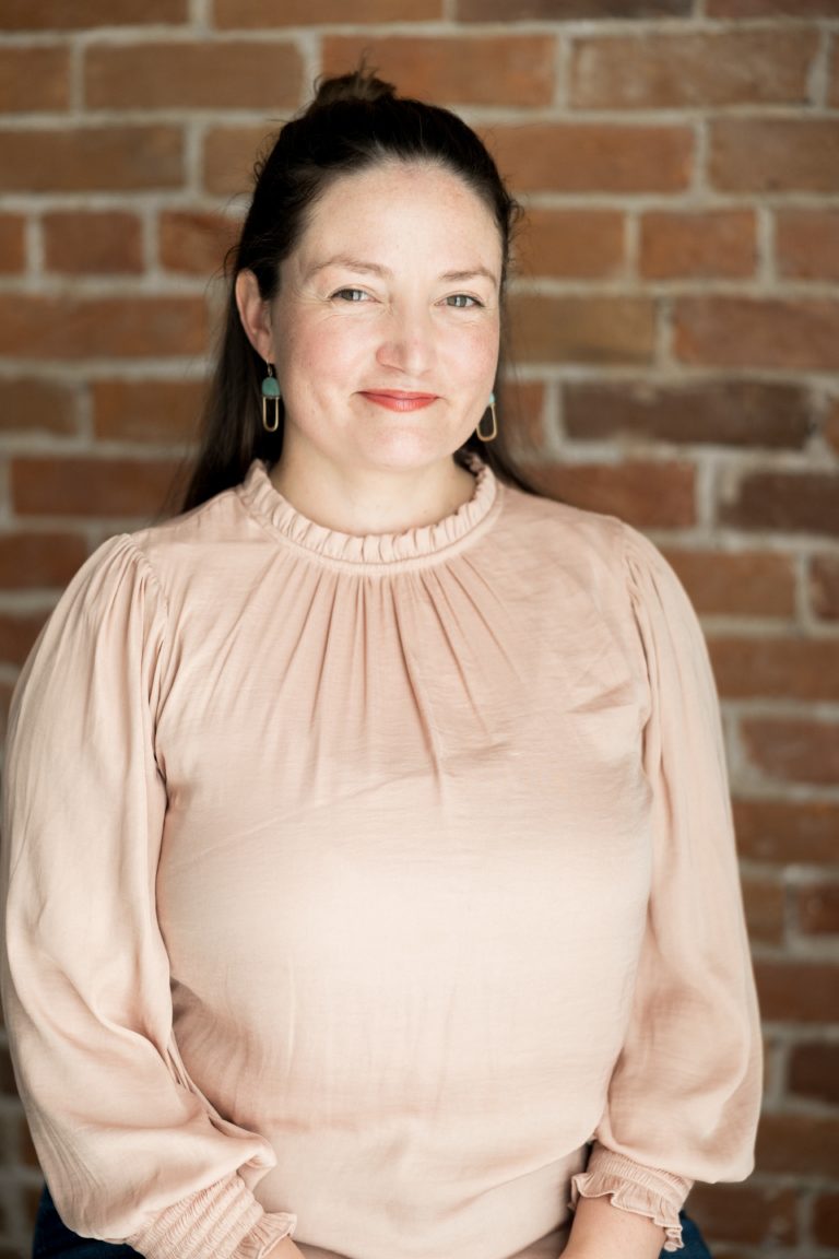 A smiling white woman with dark brown hair in a ponytail wearing a pink blouse.